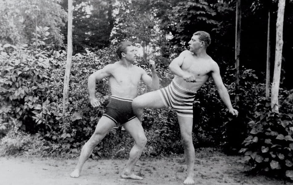 Photographie noir et blanc montrant deux hommes effectuant des prises de boxe devant une futaie. Ils ne sont vêtus que d'un short.