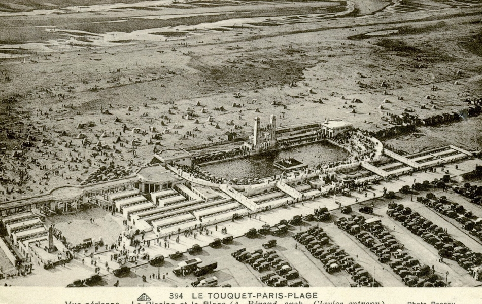 Carte postale noir et blanc aérienne montrant une plage et une piscine extérieure au bout de laquelle est accolé un parking.