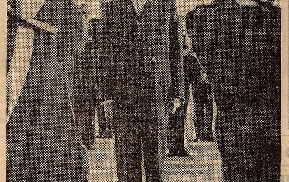 Coupure de presse montrant une photographie d'un homme debout, vêtu d'un costume, au garde-à-vous, devant le reste de la délégation. Un militaire et un porte-drapeau lui font face et l’encadrent. En-dessous, la légende suivante : "À l'arrivée du Touquet, pendant l'exécution de la Marseillaise, le général est figé au garde-à-vous".