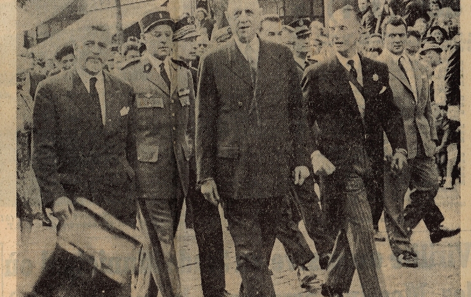 Coupure de presse illustrée d'une photographie sur laquelle on voit le général de Gaulle à pied, à la tête d’un cortège et entouré de quatre personnes : le maire de Calais et le ministre des Transports en civil, le préfet du Pas-de-Calais et l’aide de camp présidentiel en tenue d’apparat. En-dessous, la légende suivante : "Entouré de M. Buron, ministre des Travaux publics et des Transports, de son aide de camp, de MM. Cousin préfet du Pas-de-Calais et de M. Vendroux, le général de Gaulle descend le boulevard Jacquard à Calais".