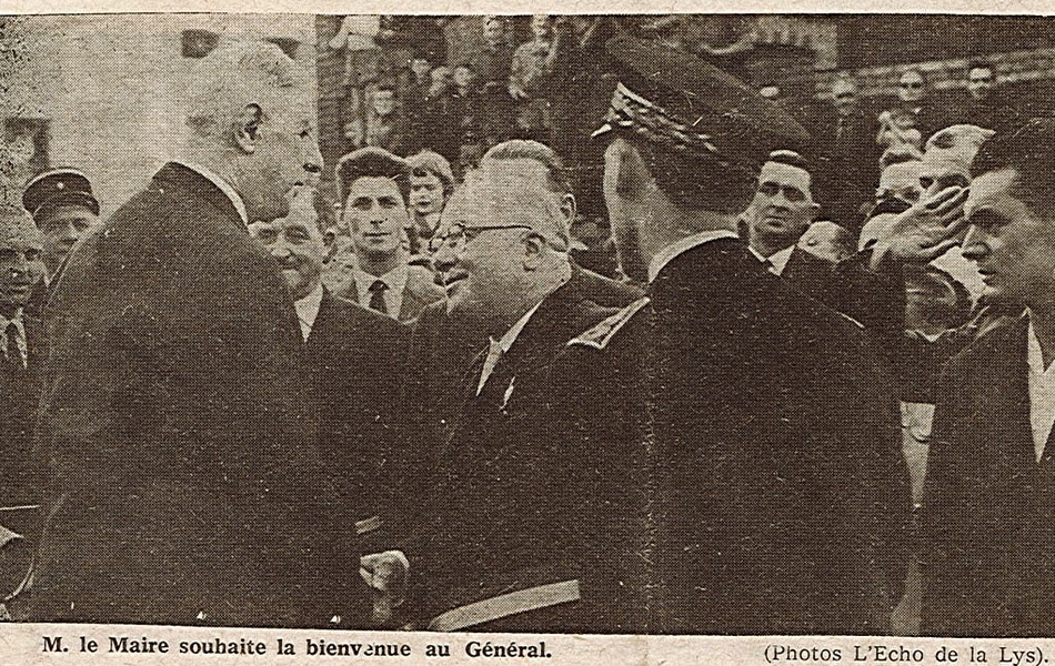 Photographie de presse sur laquelle on voit le général de Gaulle serrant la main du maire d’Aire-sur-la-Lys, devant plusieurs représentants de la commune. Le préfet du Pas-de-Calais tourne le dos au photographe. En-dessous, la légende suivante : "Monsieur le maire souhaite la bienvenue au général".