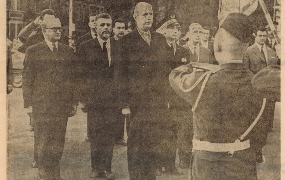 Photographie de presse sur laquelle on voit le général de Gaulle au garde-à-vous devant les membres du cortège et le maire d’Arras. Devant lui, des militaires lui présentent un drapeau.