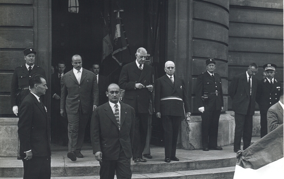 Photographie de presse sur laquelle on voit le général de Gaulle en haut des marches du perron de l’hôtel de ville, aux côtés du maire de Béthune, qui porte son écharpe tricolore à la ceinture. Ils sont entourés par des policiers en uniforme et des agents de sécurité. 