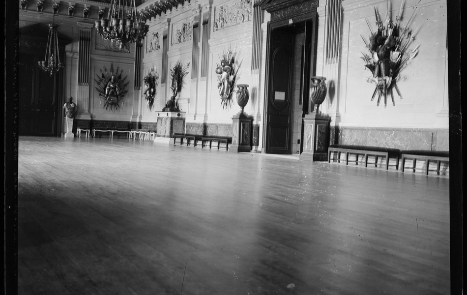 Photographie noir et blanc montrant une vaste salle vide richement ornée aux murs.