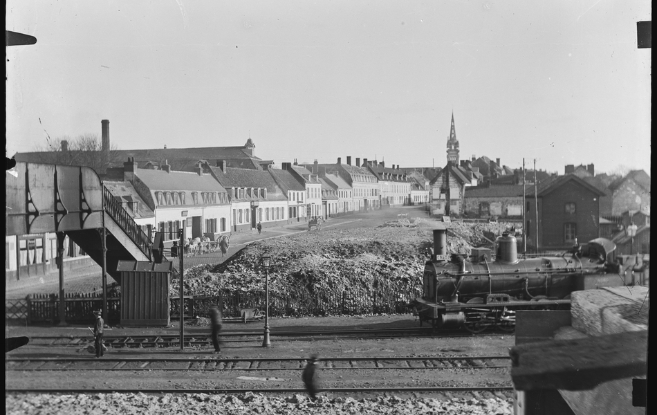 Photographie noir et blanc montrant un train à vapeur traversant une voie ferrée bordée de maisons.