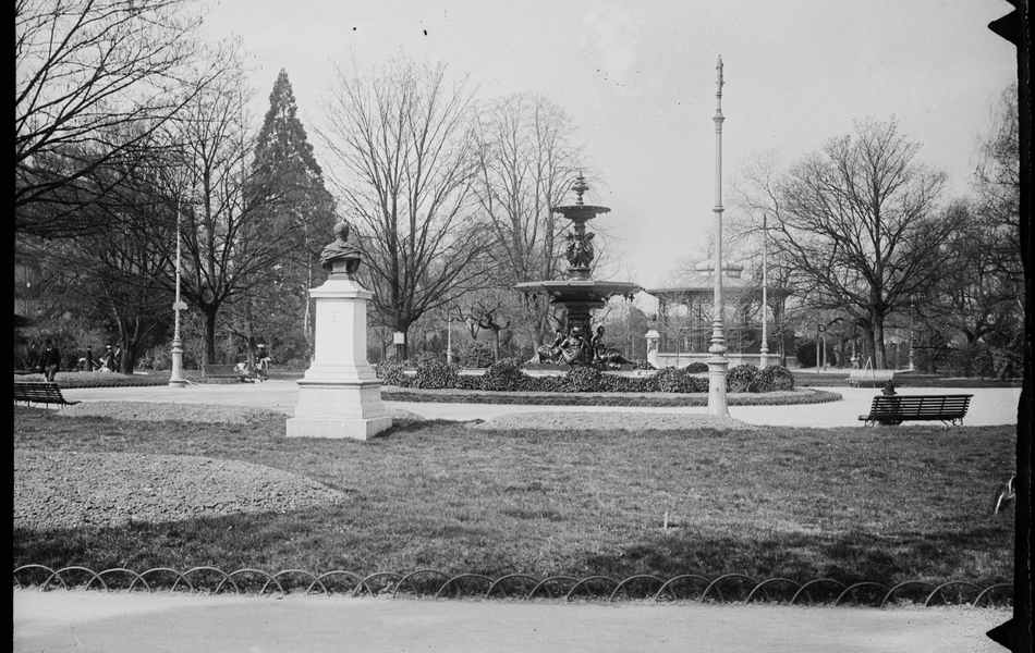 Photographie noir et blanc montrant un square avec une fontaine, un kiosque à musique et un buste en pierre.