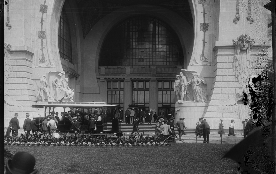 Photographie noir et blanc montrant le parvis d'un grand bâtiment devant lequel stationne un tramway.